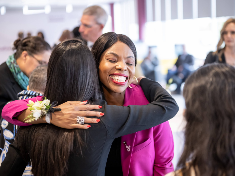 Black businesswoman in a purple blouse smiles widely while hugging a woman in a black blouse.