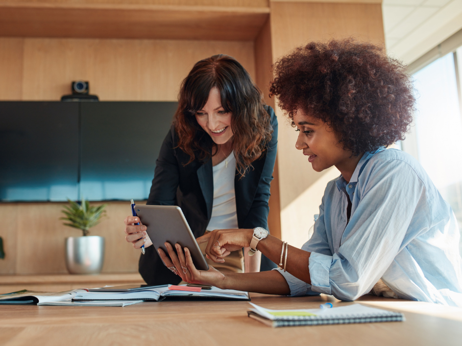 Two women in an office hold up a tablet, one pointing at the screen while the other observes.