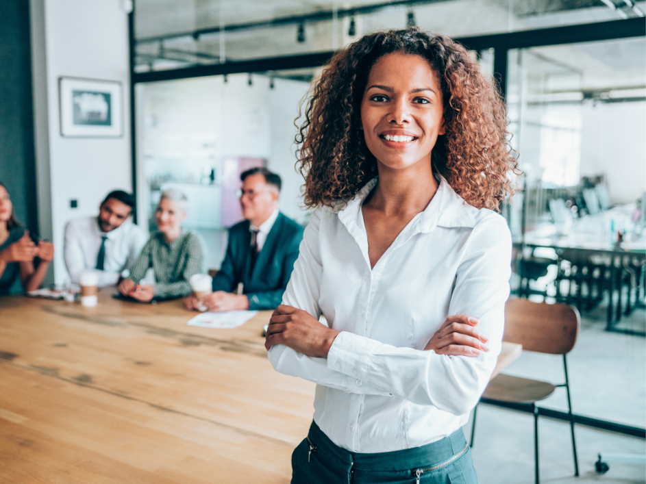 A Black woman with curly hair folds her arms and smiles while standing in a meeting room.