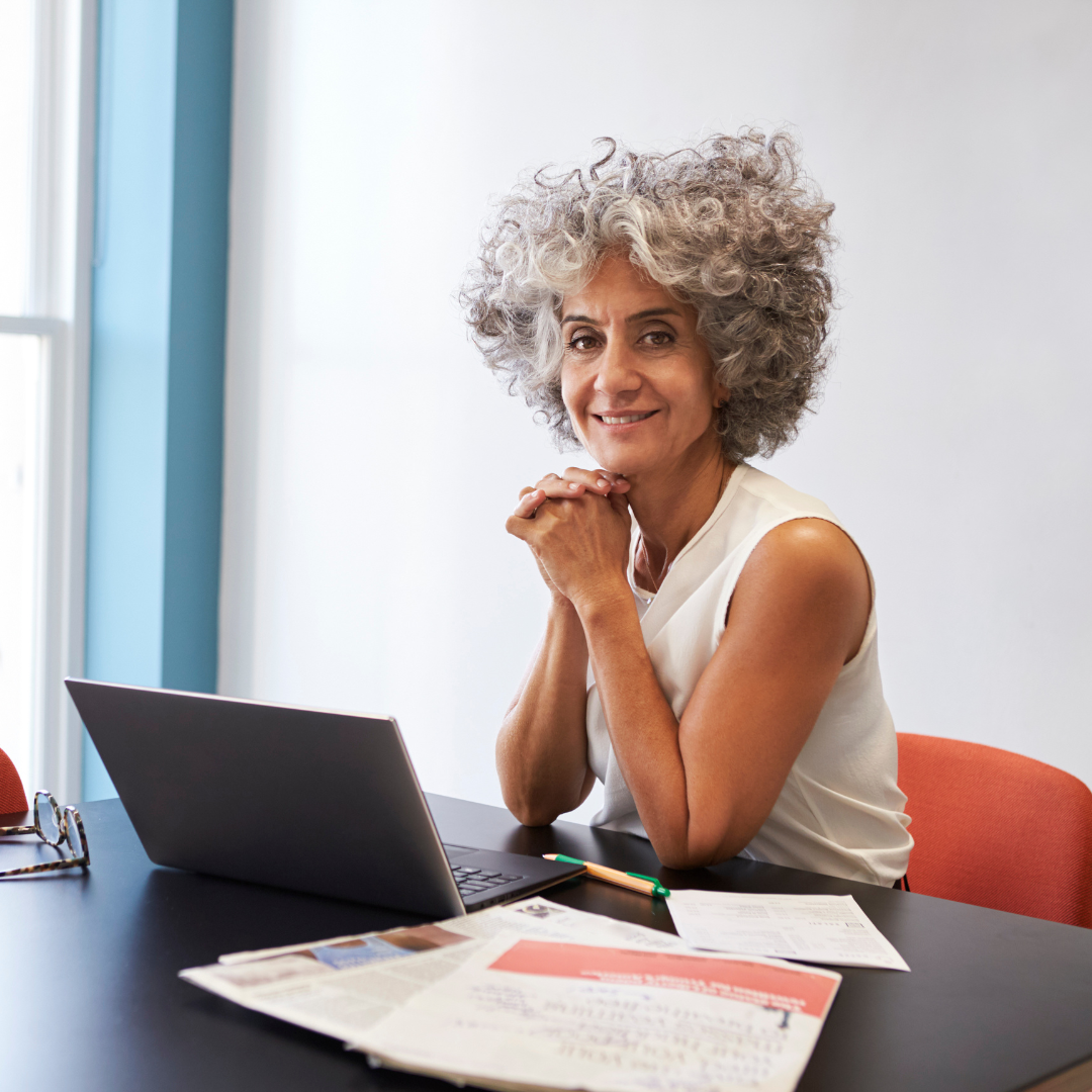 Curly-haired older White woman smiles at her desk with folded hands in front of an open laptop.