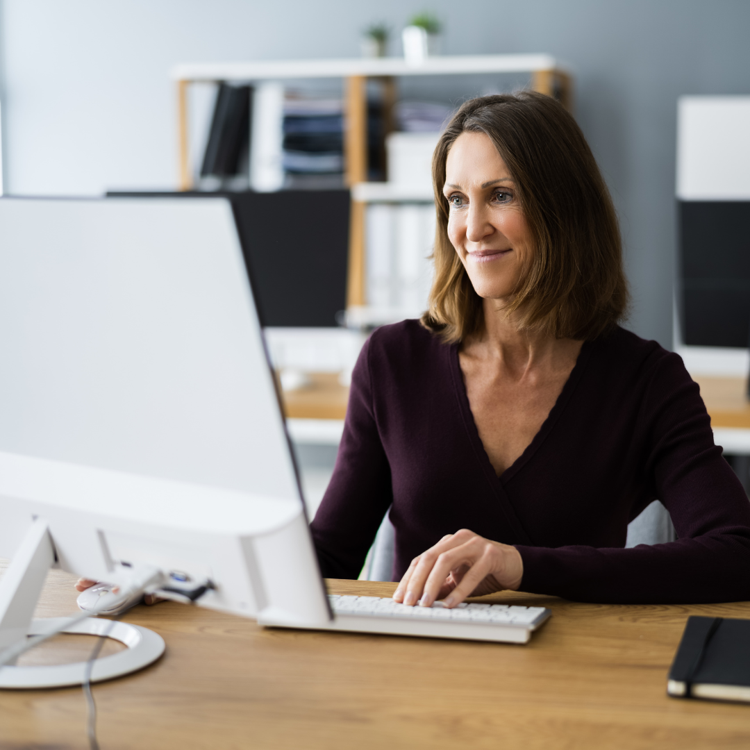 A smiling woman sitting at her office desk typing on a keyboard as looks at the screen.