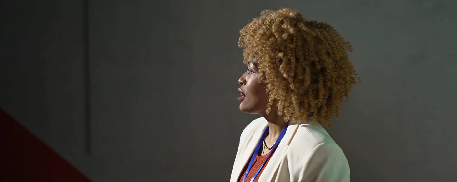 Black businesswoman speaking from a stage with blond curly hair and cream suit.
