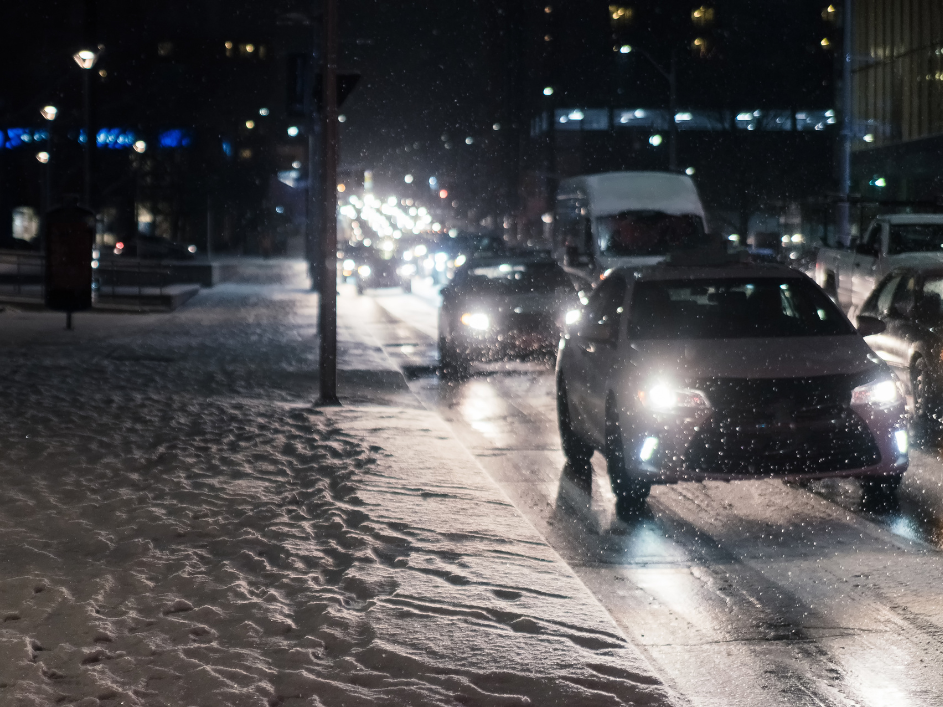 Cars driving down a street during a snowy night with snow piling up on the sidewalks and grass.